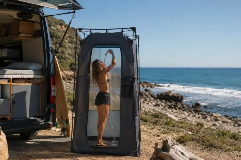 Cabine de douche amovible installée à côté d’un van aménagé en bord de mer avec une personne à l’intérieur