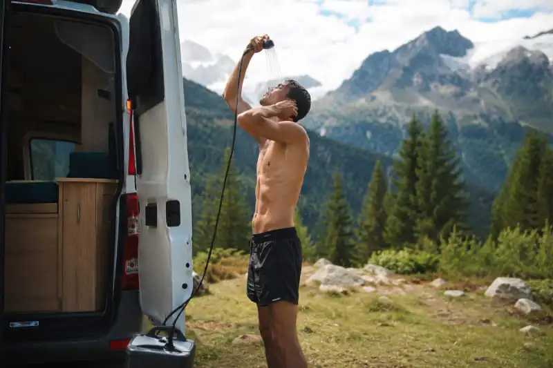Personne prenant une douche extérieure avec une douche amovible à côté d’un van aménagé en montagne
